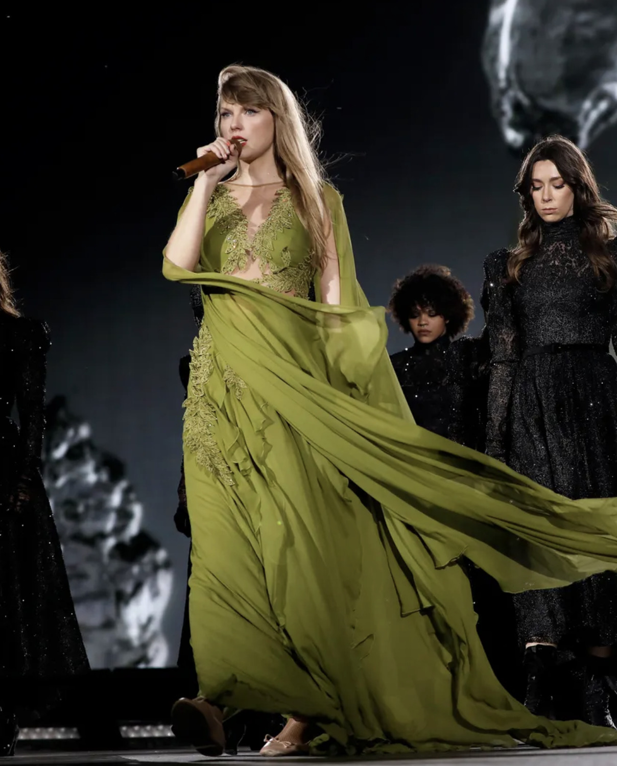 Taylor Swift performing on stage during the 'Eras Tour,' wearing a green lace gown with a flowing cape, embodying the nature-inspired and poetic aesthetic of her 'Folklore' album era.